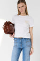 A person wearing a white t-shirt and blue jeans holds a large brown leather bag, standing against a plain white background.
