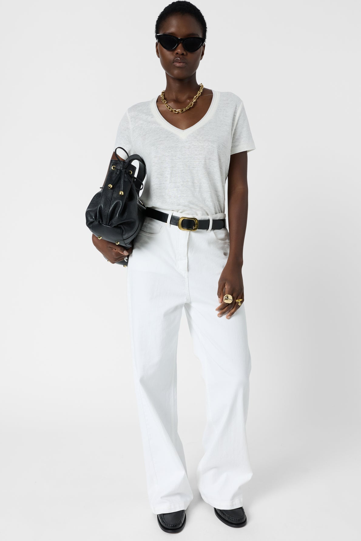 Woman wearing sunglasses, a gold necklace, a light gray t-shirt, white wide-leg pants, and a black belt, holding a black handbag. She stands confidently against a plain white background.
