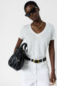 A person wearing a white V-neck T-shirt, white pants, black sunglasses, gold chain necklace, black belt, and holding a black handbag with gold accents poses against a plain light background.