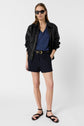 A woman stands against a white background wearing sunglasses, a black leather jacket, a blue V-neck shirt, black shorts with a belt, and black sandals. She has straight hair and a confident posture.