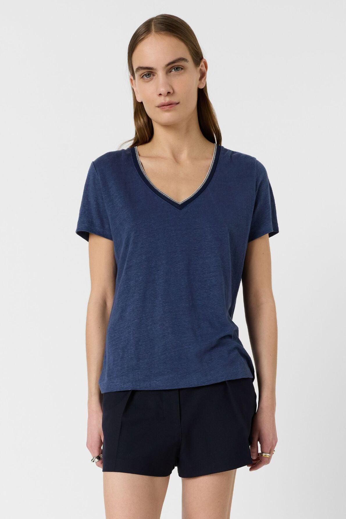 A person with long brown hair wears a blue V-neck T-shirt and black shorts, standing against a plain white background.