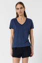 A person with long brown hair wears a blue V-neck T-shirt and black shorts, standing against a plain white background.