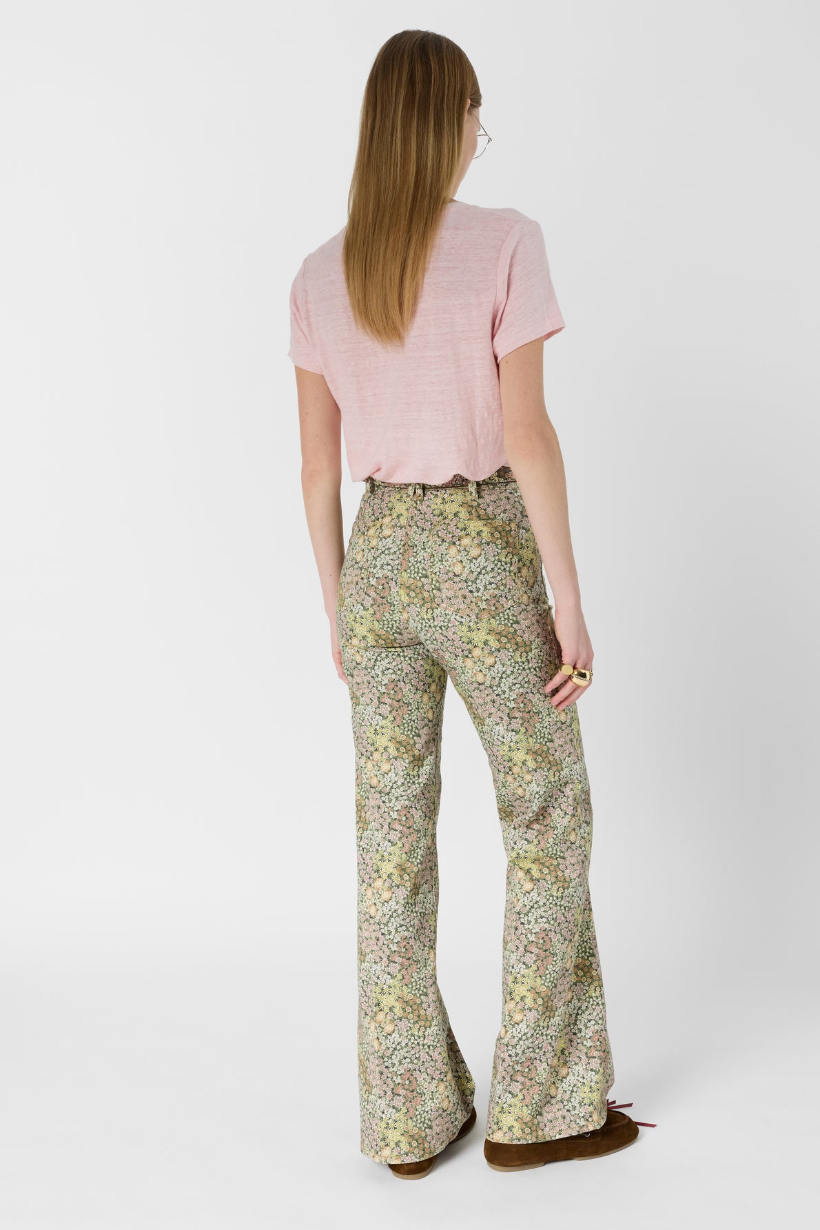 A person with long straight hair, wearing a light pink T-shirt and high-waisted, flared floral pants, stands facing away from the camera against a plain white background.