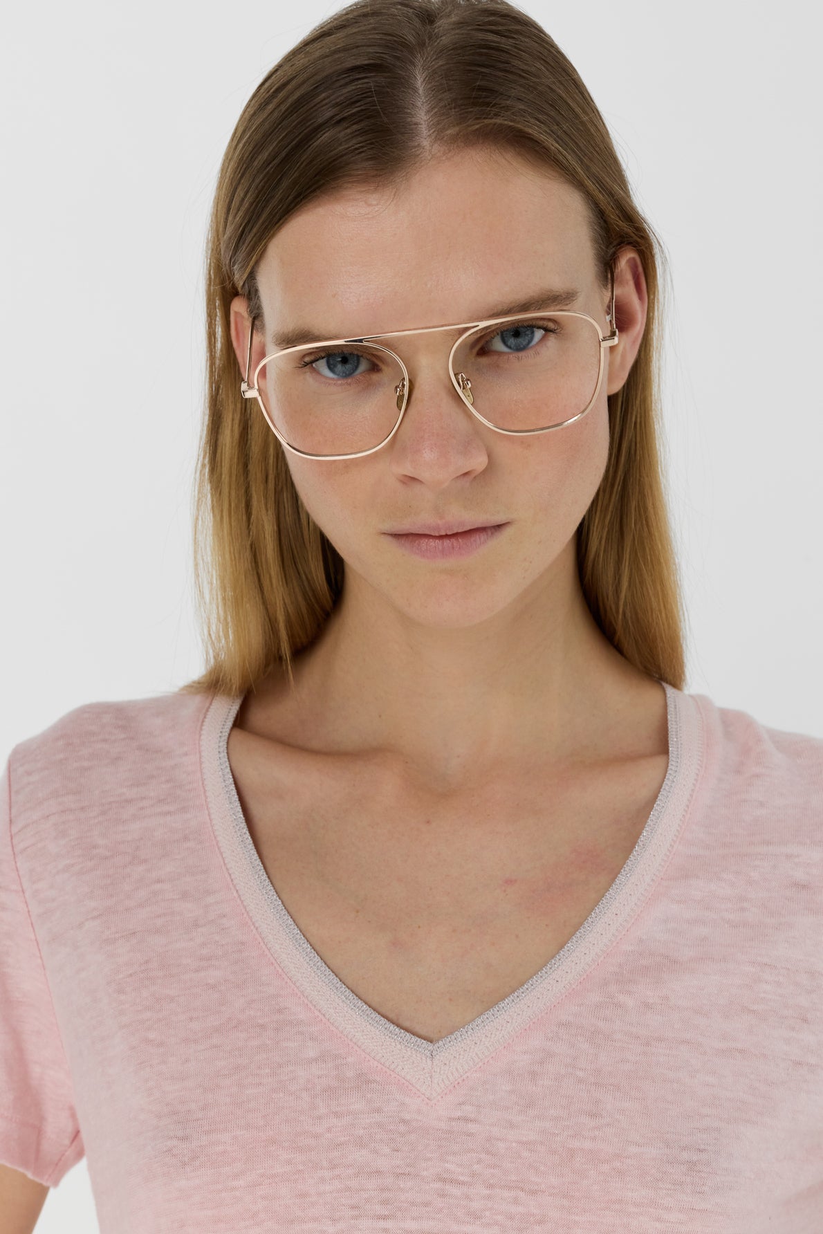 A woman with straight blonde hair wears gold-rimmed glasses and a light pink V-neck shirt, standing against a plain white background, looking directly at the camera with a neutral expression.