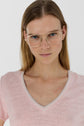 A woman with straight blonde hair wears gold-rimmed glasses and a light pink V-neck shirt, standing against a plain white background, looking directly at the camera with a neutral expression.