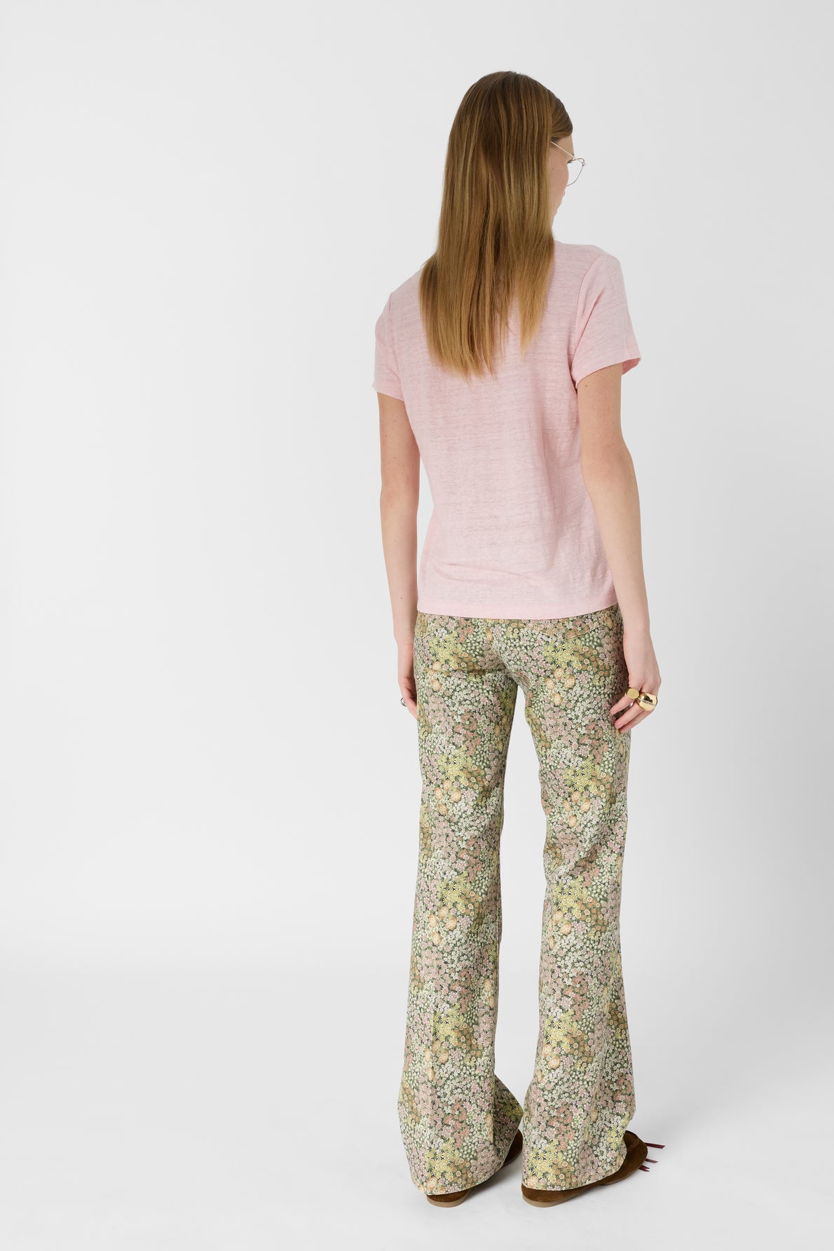 A person with long hair, wearing a light pink short-sleeve shirt and floral bell-bottom pants, stands facing away from the camera against a plain white background.