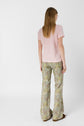 A person with long hair, wearing a light pink short-sleeve shirt and floral bell-bottom pants, stands facing away from the camera against a plain white background.