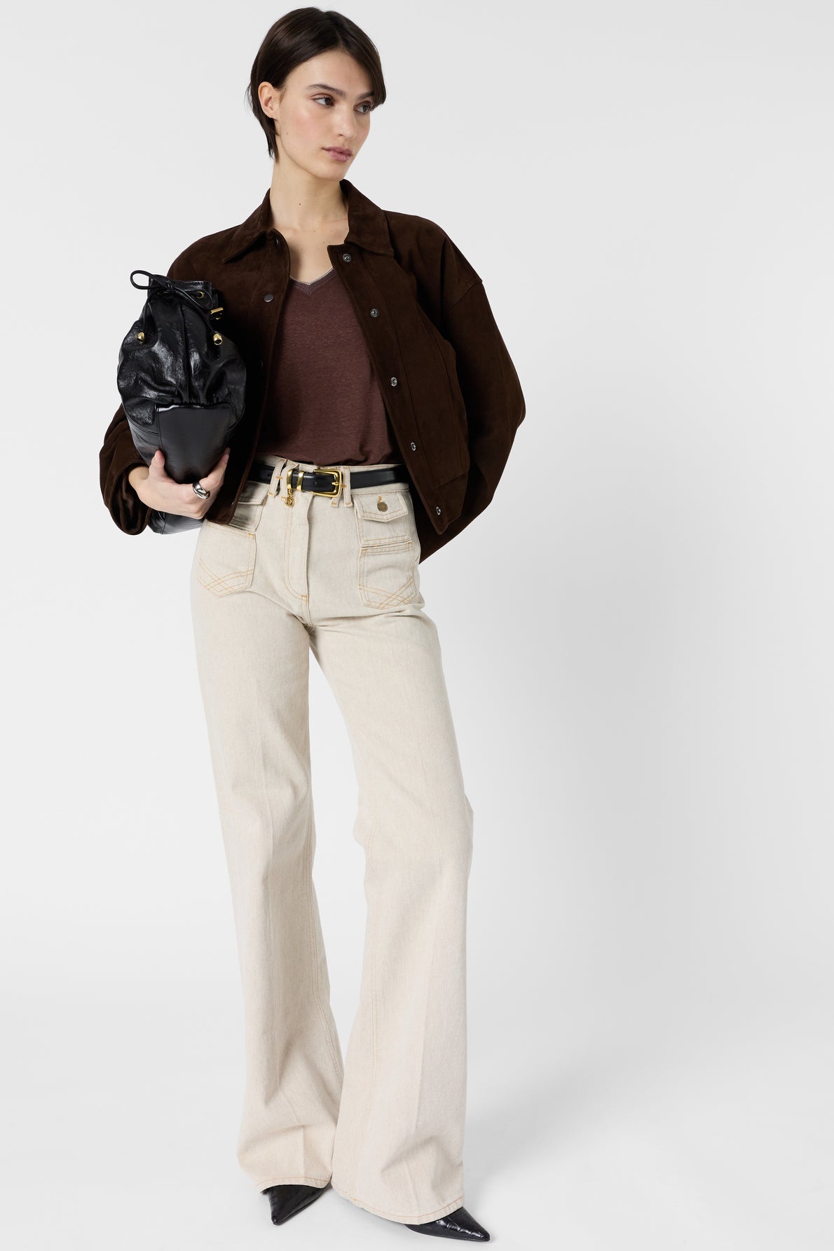 Woman standing against a white background, wearing a brown jacket, brown shirt, high-waisted beige pants, black pointed shoes, holding a black bag under her arm, looking slightly to the side.
