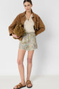 A woman stands against a plain background, wearing a brown suede jacket, beige top, floral shorts, brown sandals, and holding a brown suede bag under her arm.