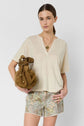 Woman with straight brown hair wears a cream top, floral shorts, and layered necklaces. She holds a brown drawstring bag and stands against a plain white background, looking confidently at the camera.
