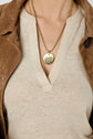 A person wearing a beige V-neck top, a brown suede jacket, and two gold necklaces, one with a round pendant.
