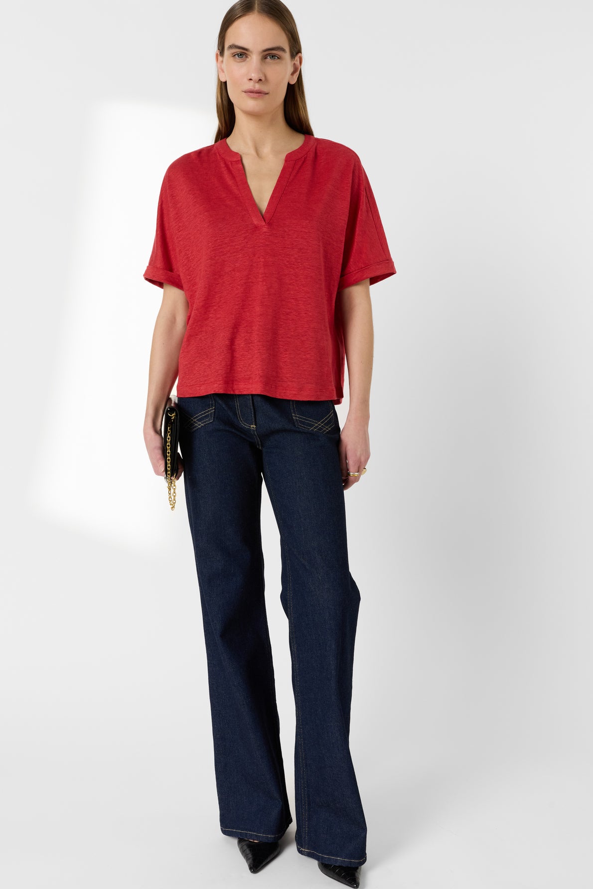 A person stands against a white background wearing a loose red V-neck short-sleeve top, dark blue bootcut jeans, and black pointed shoes, holding a black clutch with a gold chain.