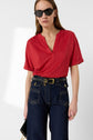 Woman wearing a red V-neck short-sleeve top, high-waisted dark blue jeans with front pockets, a black belt with a gold buckle, black sunglasses, and holding a small black handbag.