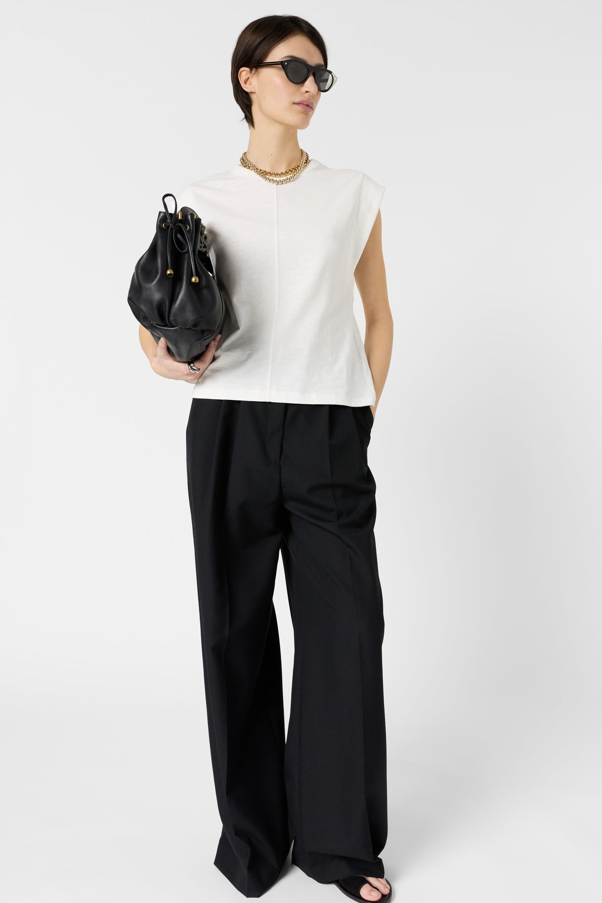 Woman wearing black sunglasses, white sleeveless top, black wide-leg pants, and sandals, holding a black handbag, stands against a plain white background.