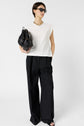Woman wearing black sunglasses, white sleeveless top, black wide-leg pants, and sandals, holding a black handbag, stands against a plain white background.