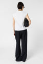 Person standing facing away from the camera, wearing a white sleeveless top, black wide-leg pants, and holding a black handbag. The background is plain white.