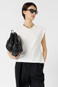 Woman with short dark hair wears sunglasses, a white sleeveless top, black pants, and a gold chain necklace, holding a large black bag against a plain white background.