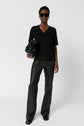 A person wearing sunglasses, a black lace-trimmed V-neck top, black leather pants, and black shoes holds a black handbag, standing against a plain white background.
