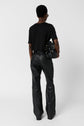Person standing against a white background, facing away. They wear a black short-sleeve shirt, black leather pants, black shoes, and hold a shiny black handbag in their left hand.