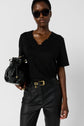 A person wearing black sunglasses, a black lace-trimmed top, and black leather pants holds a black leather bag with gold accents against a plain background.