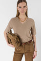 A person wearing a beige t-shirt en V and brown pants holds a brown suede handbag. They have straight, shoulder-length hair and wear a gold chain necklace, bracelet, and a brown belt, standing against a plain white background.