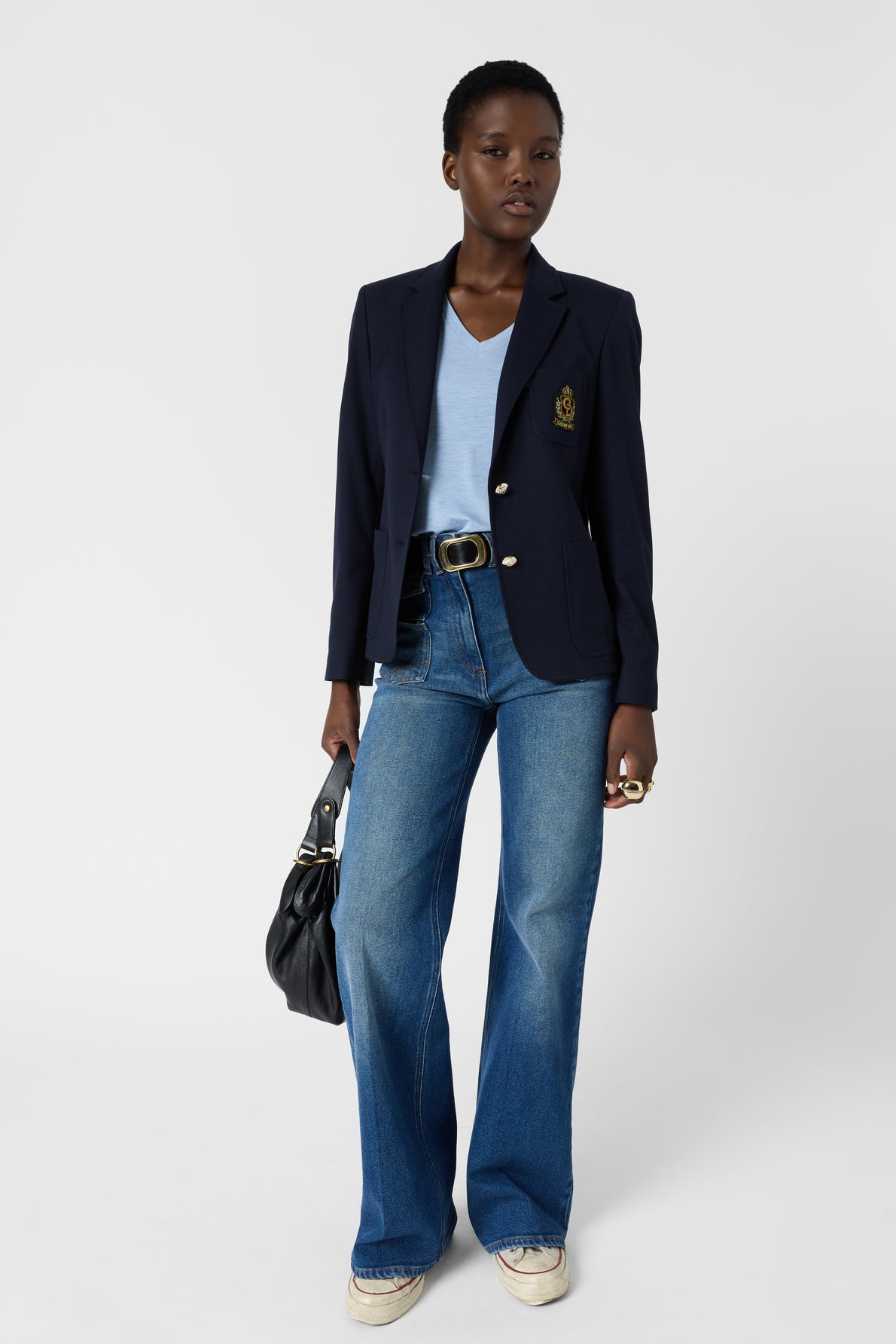 A person stands wearing a navy blazer over a bleu ciel t-shirt en V, paired with wide-leg jeans and sneakers for a look casual. They hold a black handbag and wear rings, with short curly hair and a confident expression against a plain white background.