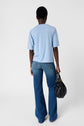 A person stands facing away, wearing a bleu ciel t-shirt en V and blue flared jeans, holding a black handbag in their right hand against a plain white background for a chic yet look casual.