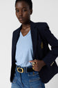 A person wearing a navy blazer with a crest, a bleu ciel t-shirt en V, blue jeans, a black belt with a gold buckle, and several gold rings poses with one hand on their hip against a plain background for a look casual yet refined.
