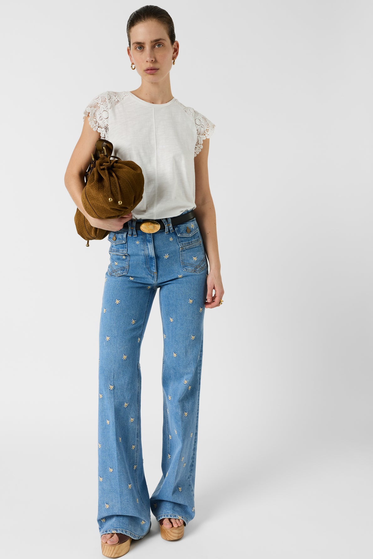 Woman stands against a white background wearing a white lace-sleeved blouse, high-waisted blue jeans with small embroidered designs, tan platform sandals, and holding a brown handbag.