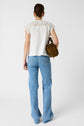 Woman standing with her back to the camera, wearing a white lace-trimmed top and blue jeans with small floral patterns. She holds a brown handbag and has her hair tied back. Background is plain and white.