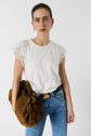 Woman wearing a white lace-sleeve top and blue embroidered jeans, holding a large brown bag, standing against a plain white background.