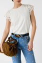 A woman wearing a white lace-sleeve top and blue jeans with embroidered stars holds a brown textured handbag. Only her upper body is visible against a plain background.