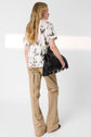 A person with long blonde hair, wearing a white t-shirt with butterfly prints and beige flared pants, stands sideways holding a large black fringe bag against a plain background.