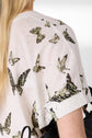 A person with long blonde hair wears a white T-shirt patterned with green and black butterflies. The photo focuses on the shirts sleeve and shoulder. The person holds a black bag over their arm.