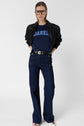 A person stands against a white background wearing glasses, a black leather jacket, a tee-shirt bleu marine en coton Darel, high-waisted dark blue jeans, and a black belt with a gold buckle.
