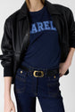 Person wearing a black leather jacket, navy blue coton tee-shirt bleu marine with AREL in bold blue letters, dark denim jeans, and a black belt with a gold buckle. The head is partially out of frame; the look embodies Darel’s effortless modern style.