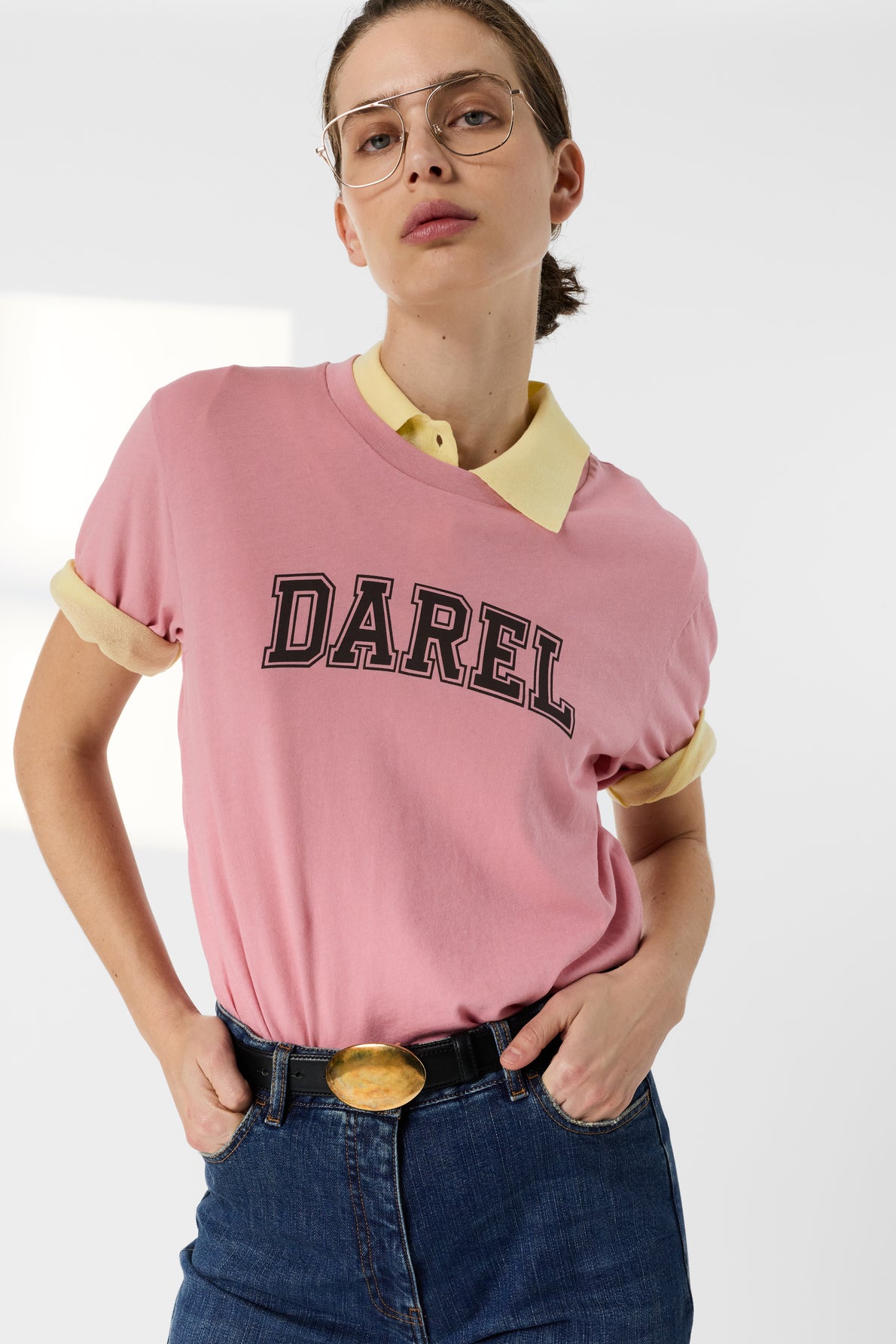 Woman wearing glasses, a tee-shirt rose en coton with the inscription Darel, a yellow collared shirt underneath, and blue jeans with a black belt, standing with hands on hips against a light background.