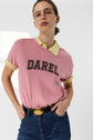 A person wearing glasses, a pink tee-shirt rose in soft coton with the inscription Darel over a yellow collared shirt, and blue jeans stands with hands on hips against a bright, minimal background.