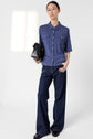 Person wearing glasses, a bleu indigo t-shirt col polo in maille douce, wide-leg dark jeans, and black shoes holds a black handbag. They stand against a plain white background, looking at the camera.