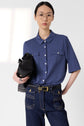 A person with long dark hair, wearing glasses, a bleu indigo t-shirt col polo, and high-waisted jeans holds a black bag against a plain light background.