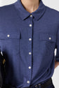A person wears a bleu indigo button-up shirt with two chest pockets and white buttons, paired with dark jeans. The photo is cropped to show only the torso.
