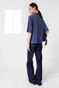 A person with long dark hair, wearing glasses, a loose bleu indigo t-shirt col polo in maille douce and dark blue jeans stands facing away, holding a black bag over one shoulder in a minimal, bright room.