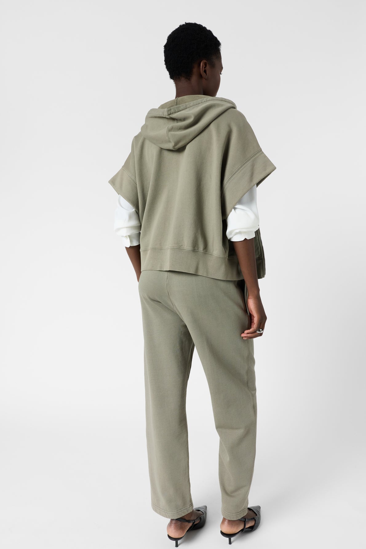 Person standing with back facing camera, wearing an olive green sweat à capuche oversize vest, matching pants in coton épais, a white long-sleeve shirt, and black sandals against a plain light background.