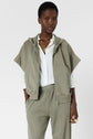 A person wears a light olive zip-up sweat à capuche oversize manches courtes, crafted from coton épais, over a white button-up shirt, matching joggers, gold necklaces, and a ring, standing against a plain white background.