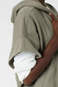 A person in an olive green sweat à capuche oversize manches courtes, crafted from coton épais and layered over a white long-sleeve shirt, stands with a hand resting on their chest against a plain background.
