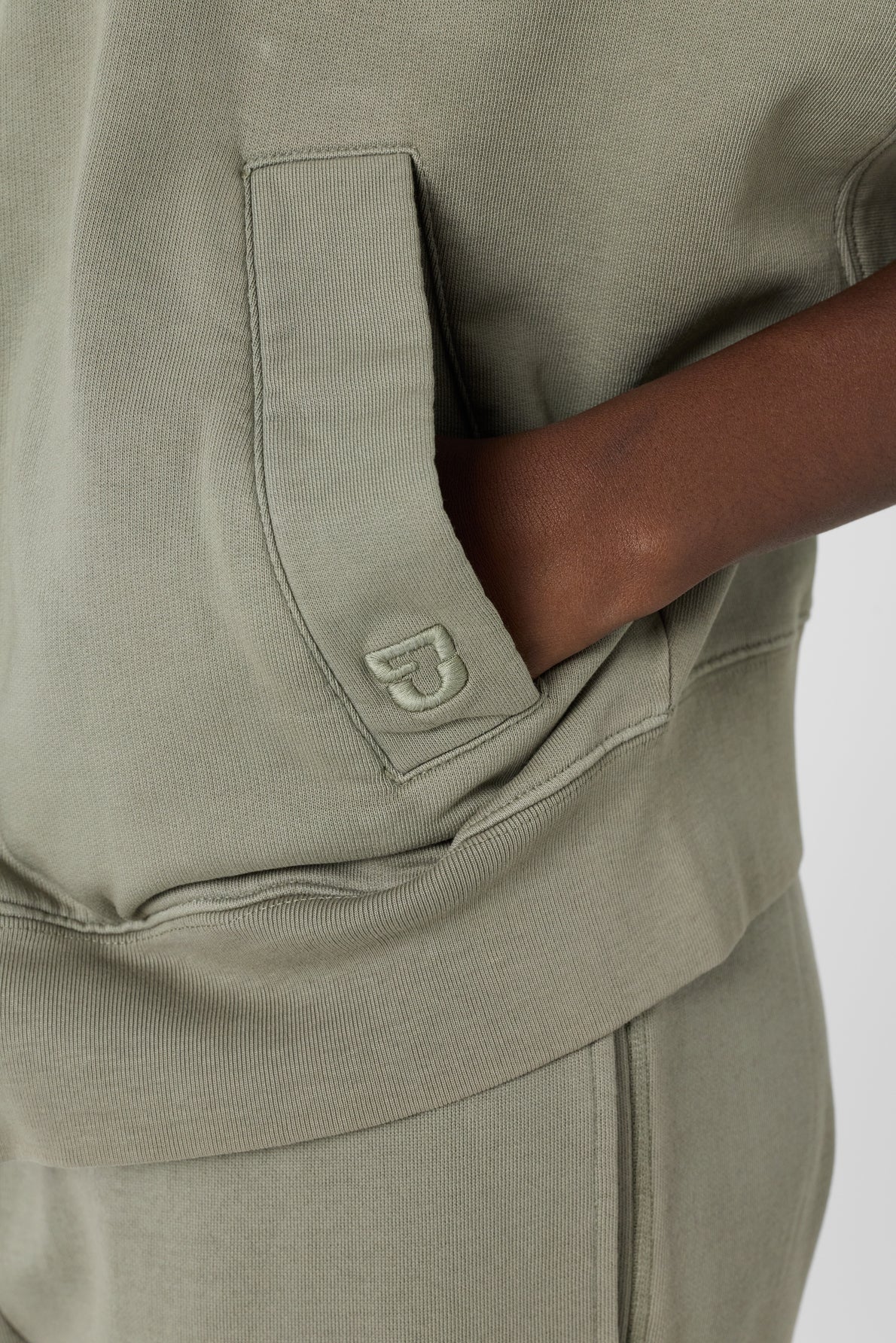 A person wearing an olive green tracksuit with one hand in the pocket. Crafted from coton épais, the sweat à capuche oversize features a small embroidered B logo on the pocket.