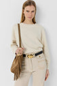Woman with straight blonde hair wears a sweat écru, beige jeans, a black belt with a gold oval buckle, and carries a brown suede fringed bag over her shoulder against a plain background.