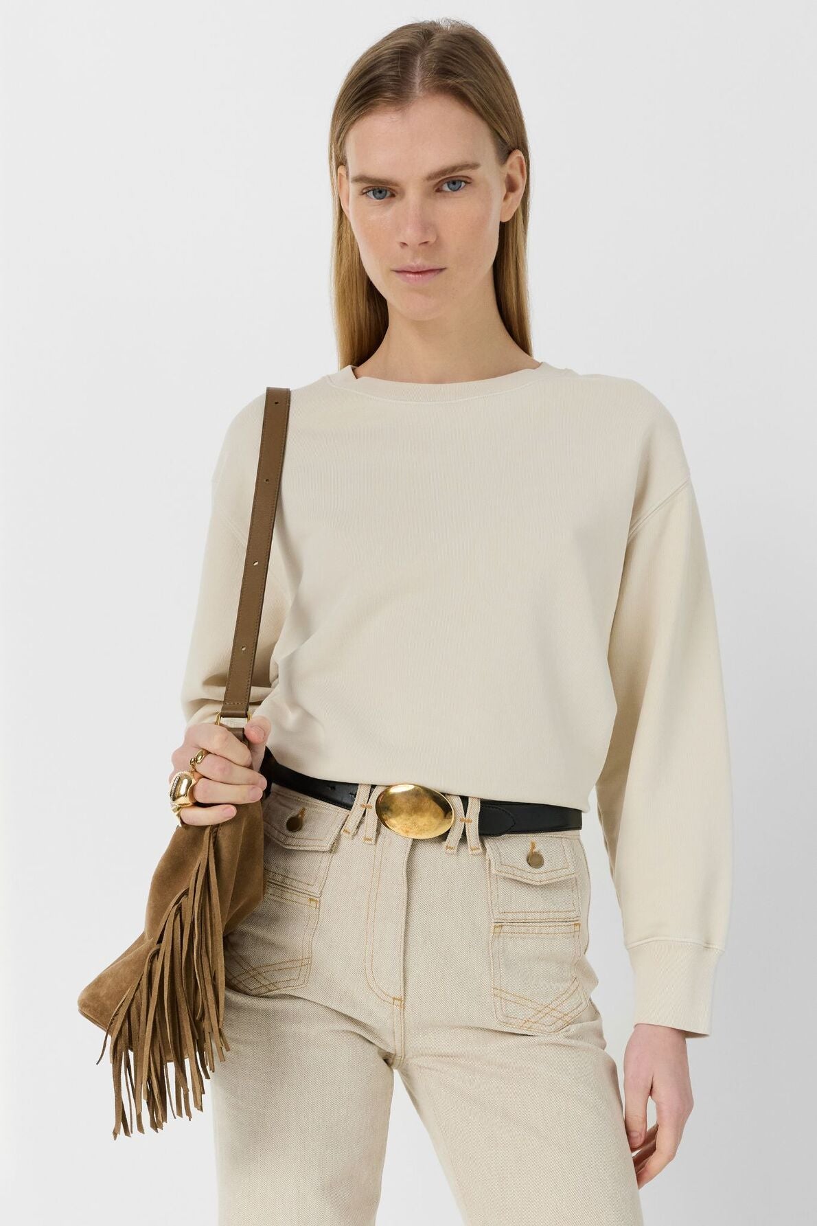 A person with straight, light brown hair wears a sweat écru and high-waisted beige jeans, holding a brown fringed bag over one shoulder, standing against a white background.