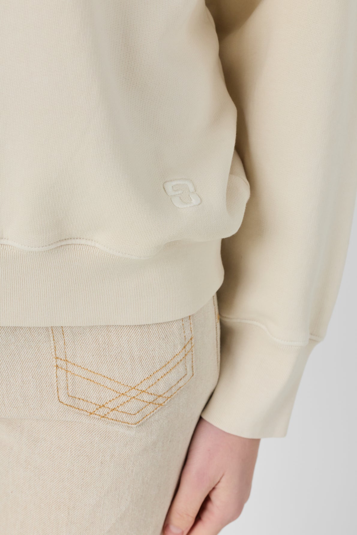 A person wearing a beige long-sleeve top, resembling an élégant sweat écru with a small embroidered logo near the hem, paired with light-colored jeans featuring visible stitching, shown from the waist down.