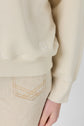 A person wearing a beige long-sleeve top, resembling an élégant sweat écru with a small embroidered logo near the hem, paired with light-colored jeans featuring visible stitching, shown from the waist down.
